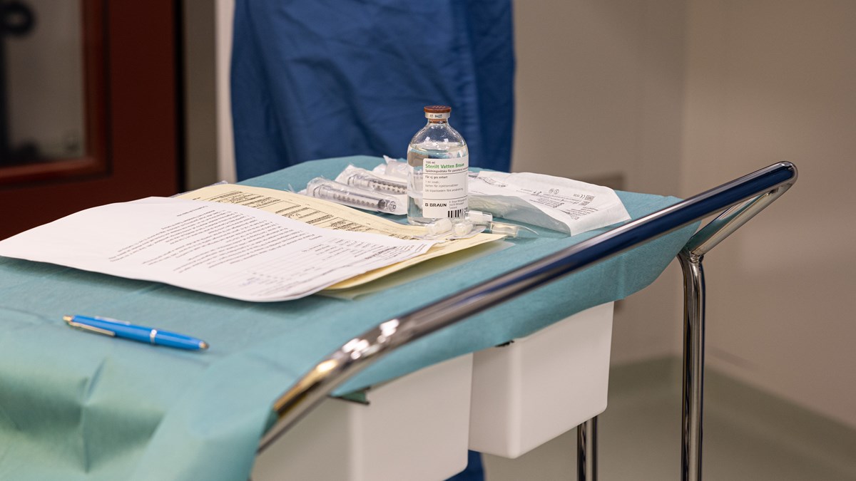  En stålvagn på hjul står i ett rum på ett sjukhus. Vagnen är täckt med blått papper, på vagnen ligger det A4 papper med text, en bläckpenna, en genomskinlig glasflaska med etikett och bredvid ligger det sprutor som är inplastade. I bakgrunden syns en person i blå läkarrock.