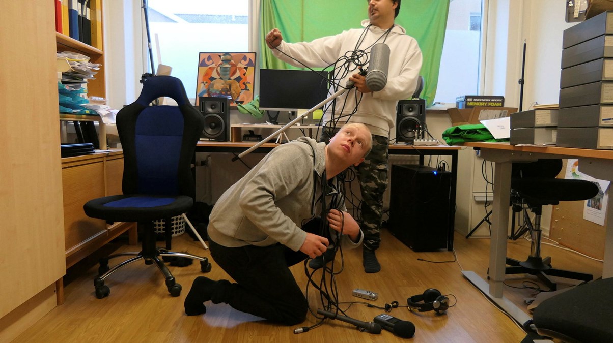 Två killar i en radiostudio.En sitter på huk på golvet, den andre står upp. De håller i mikrofoner och kablar. Vi ser hörlurar på golvet. Bilden andas lite humor.