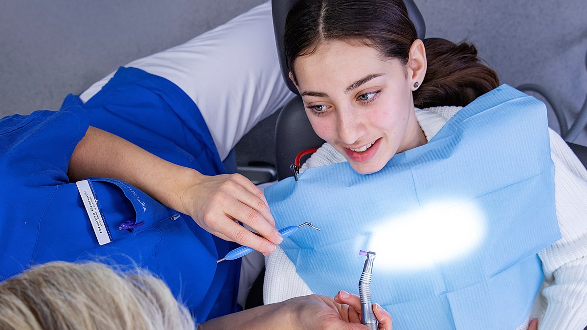 En ung kvinna med mörkt hår sitter i en tandläkarstol bredvid en kortklippt tandhygienist som håller i ett redskap.