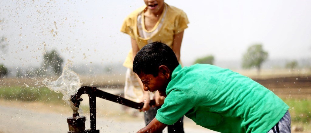 Två barn leker vid en vattenpump, vattnet sprutar och barnen skrattar.