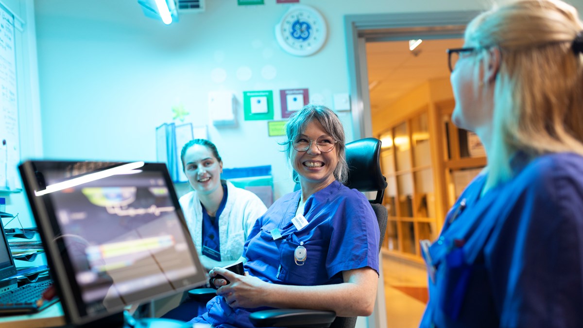 Tre medarbetare i blå sjukvårdskläder sitter vid datorskärm och samtalar leende.