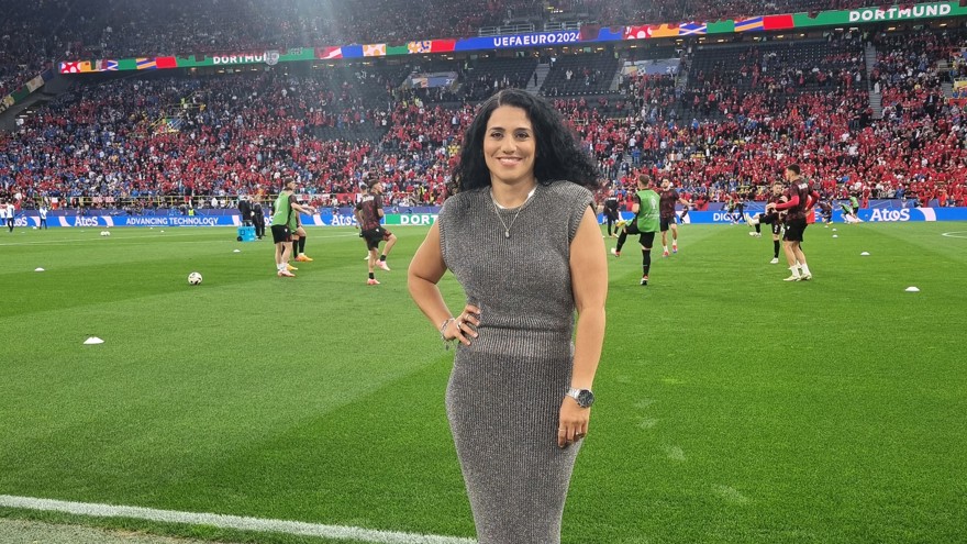 Reyhaneh Rouhani står i utkanten på en fotbollsplan. Hon har långt mörkt lockigt hår och är klädd i en grå kortärmad klänning som ser stickad ut. Hon ler mot fotografen . Bakom henne värmer fotbollspelare upp på planen, läktarna är fullsatta med mestadels rödklädda fans.