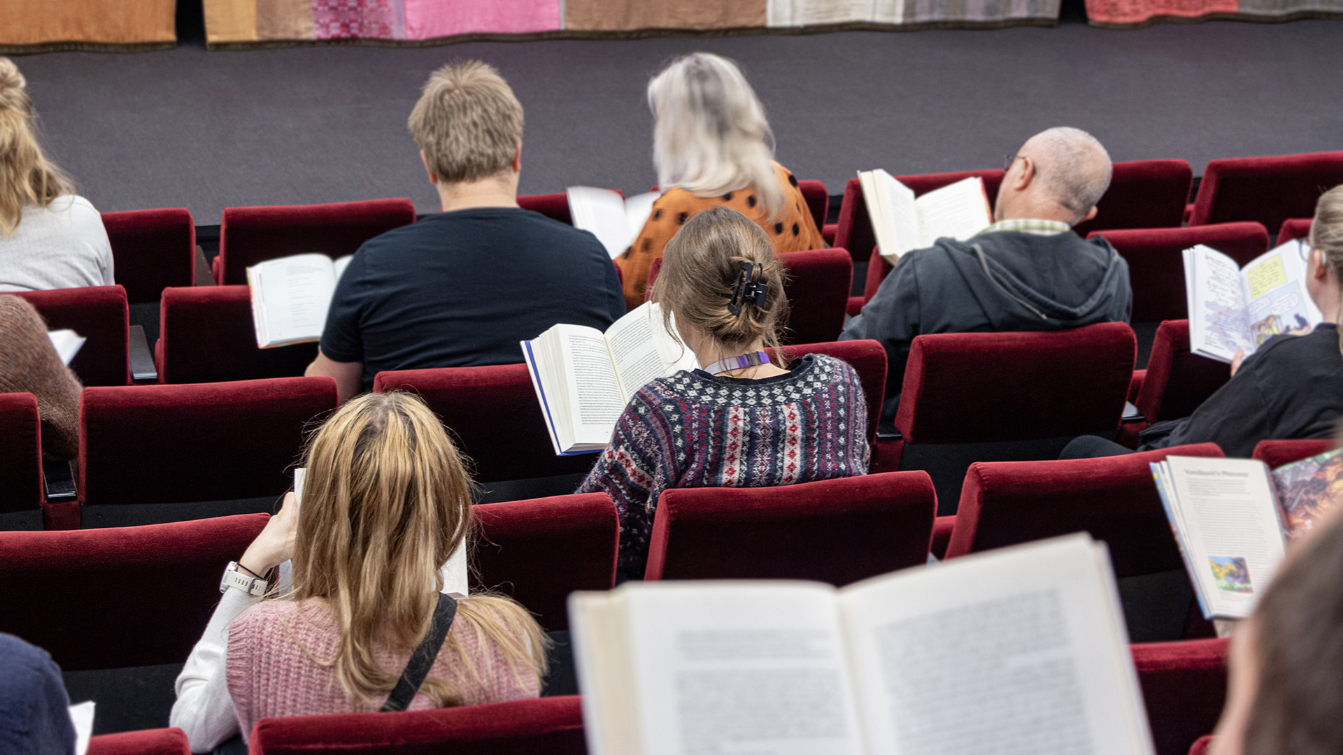 En sal med rader med röda sittplatser, som i en biosalong. Utspridda i rummet sitter människor med varsin bok uppslagen och läser.