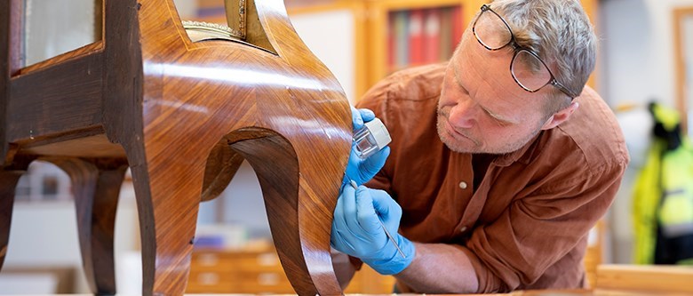 En man arbetar med ett litet verktyg och en lupp på ett träföremål. Han har ljusblå latexhandskar och glasögonen uppskjutna i pannan.