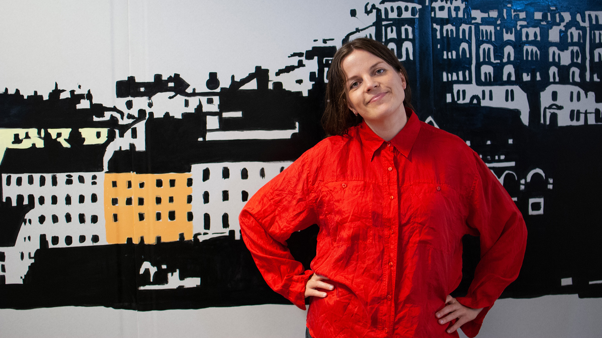 Milla Leskinen är i 35-40-årsåldern. Hon har mörkt hår som är samlat i en tofs och är klädd i en klarröd skjorta. Hon står mot en vägg med ett målat stadsmotiv, hon har händerna i midjan, huvudet lite på sned och ler med stängd mun. 