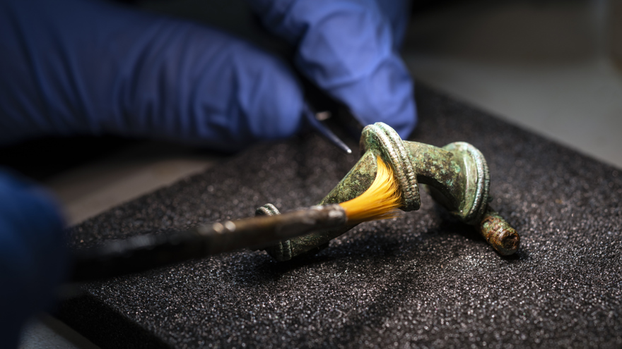 Ett mycket litet föremål av metall vilar på ett grått material. Ett par händer i ljusblå latexhandskar penslar försiktigt på föremålet.