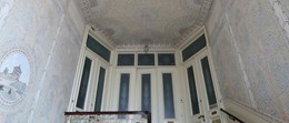 Ett äldre trapphus med målat tak och väggar, trappräcken i trä och vita dörrar med slipade glas.