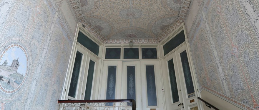 Ett äldre trapphus med målat tak och väggar, trappräcken i trä och vita dörrar med slipade glas.