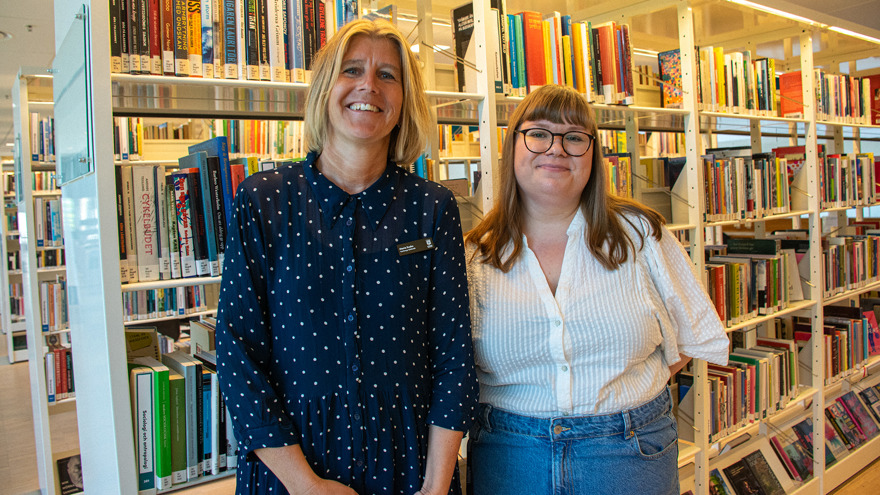Maria Svahn och Julia Brühl står framför en bokhylla inne på Varbergs stadsbibliotek. Maria har blont rakt hår till axlarna och är klädd i en blå blus med vita prickar. Julia har brunt långt hår och lugg och är klädd i jeans och en vit blus. Båda två ler mot fotografen.