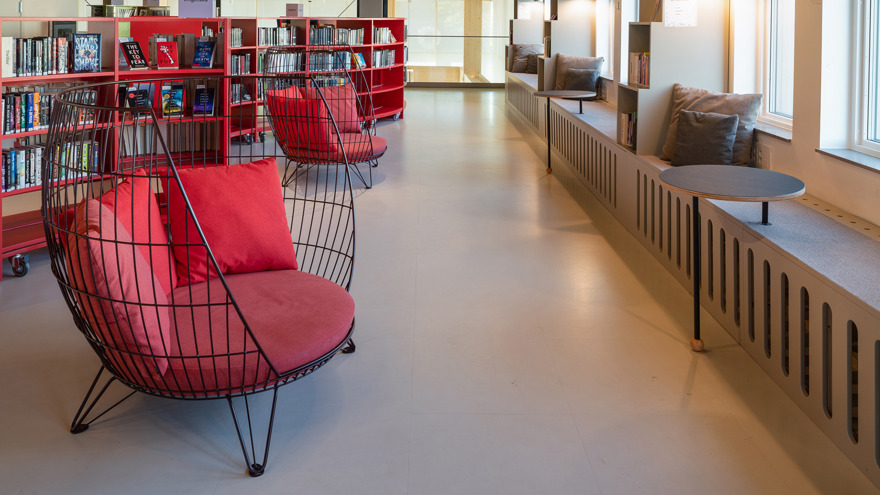 Interiör från Sollentuna bibliotek. Röda låga bokhyllor på hjul står till vänster i bild, framför dem står två stora läsfåtöljer med röda sittdynor och kuddar. Till höger finns en vägg med fönster och sittbänkar i fönstren.