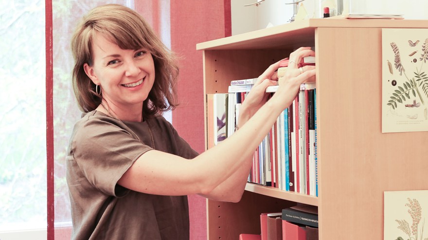 Kvinna som plockar med böcker i en bokhylla och ler mot kameran