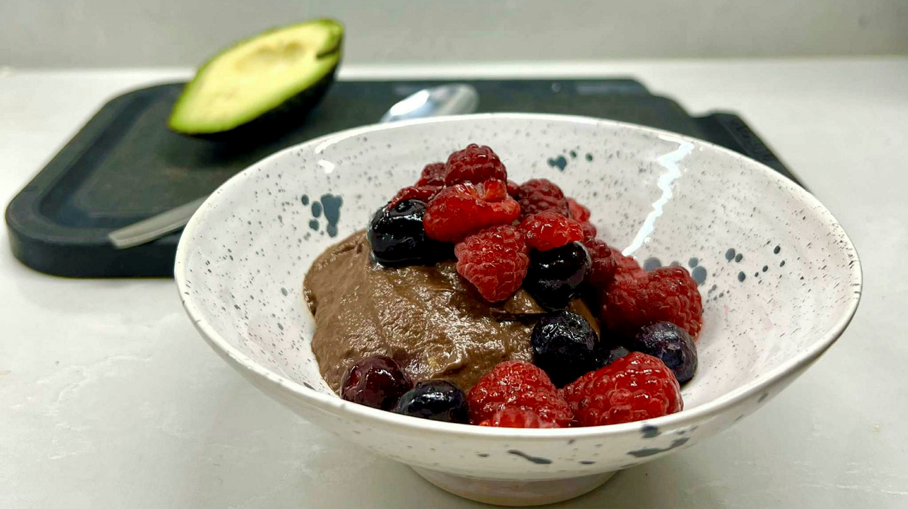 Chokladmousse i en vit skål med bär och en avokadohalva i bakgrunden