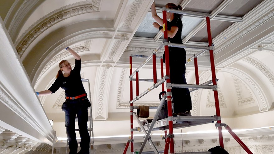 Två personer står högt uppe under ett tak och arbetar. Den ena på en stege, den andra på en byggställning.
