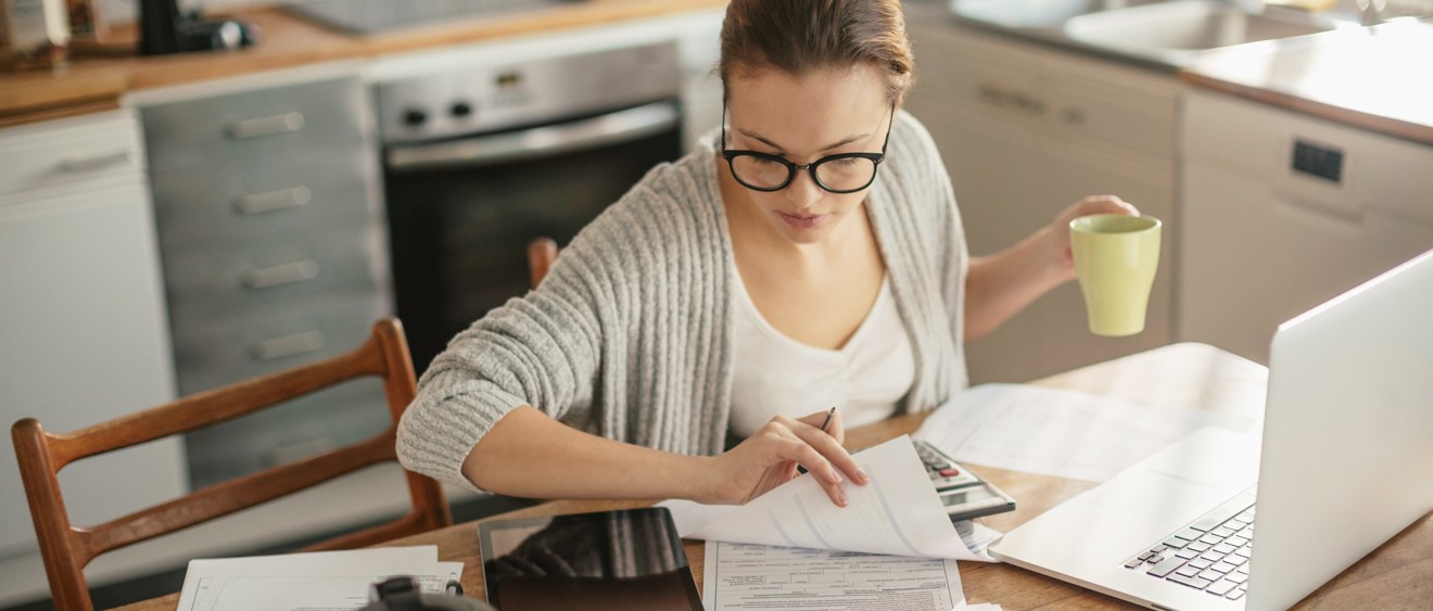 Halvbild på en person som sitter hemma i sitt kök och jobbar. Personen sitter framför en laptop, håller i en kaffekopp och har många papper framför sig.