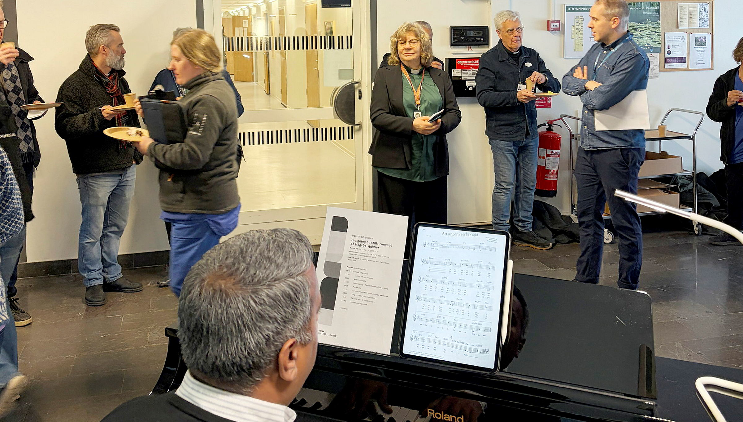 En bild som visar männsikor i ett rum som lyssnar på musik. En man spelar piano.