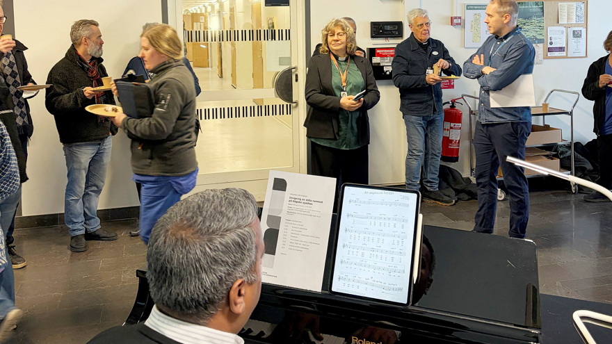 En bild som visar männsikor i ett rum som lyssnar på musik. En man spelar piano.