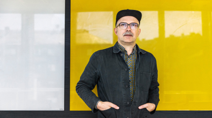 Joakim Albrektsson står framför en gulfärgad glasruta. Han 'r klädd i en svart jacka och en svart keps. Han bär glasögon och har mustasch.