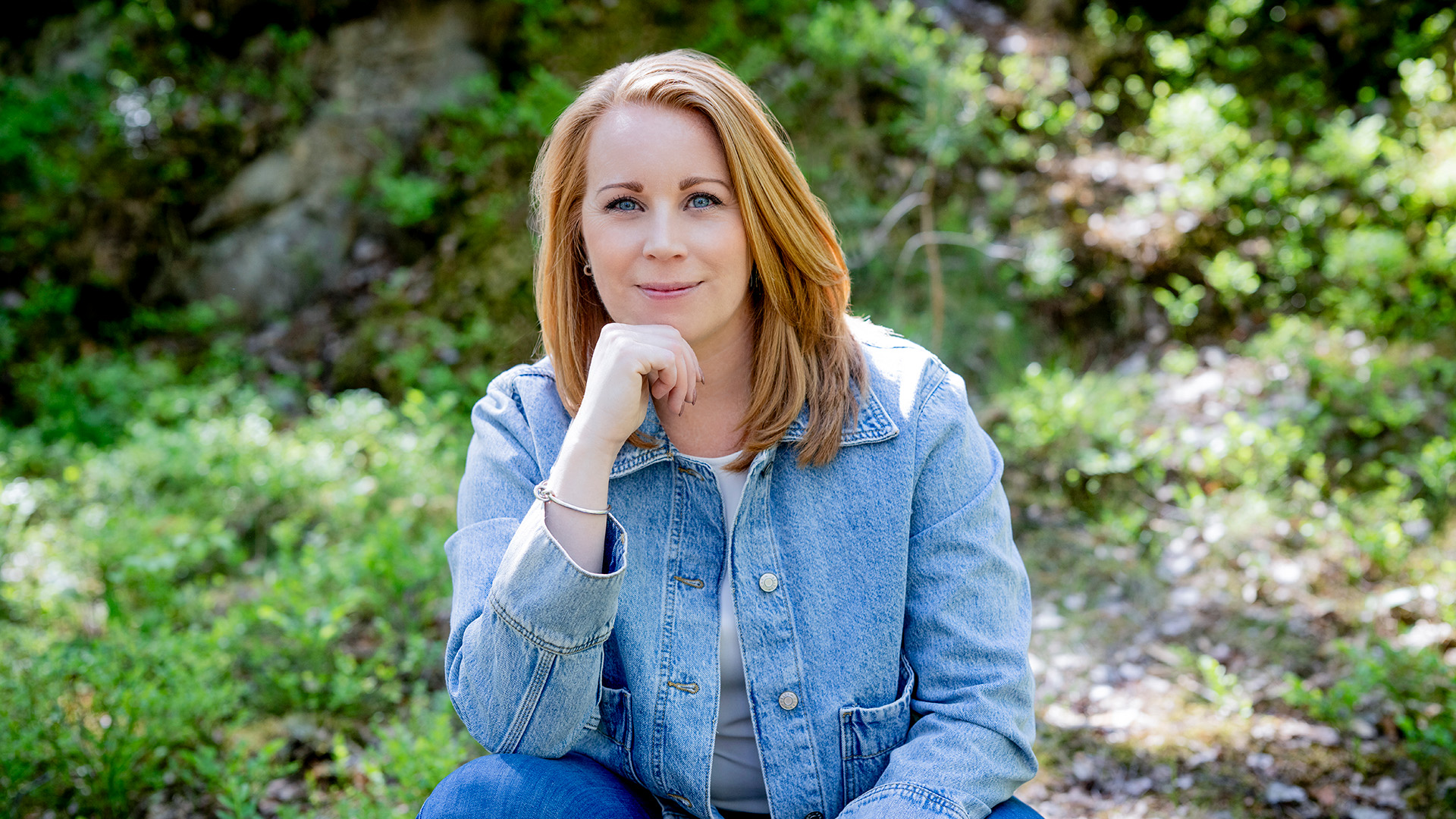 Annie Lööf sitter utomhus i en skogsbacke. Solen skiner och det är grönska bakom henne. Hon är klädd i blå jeans och en blå jacka av jeanstyg. Hon har rödblont axellångt hår och blå ögon. Hon sitter på huk och tittar rakt mot fotografen och ler med stängd mun. 