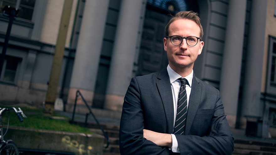 Man framför pampig byggnad. Grå kostym, vit skjorta, svart och vitrandig slips, kort hår, glasögon. Armarna i kors.