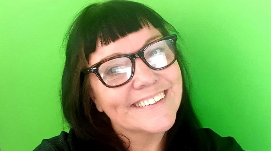 Kvinna. LÅngt svart hår. Lugg, svarta glasögon, svart blus. Hon står framför en grön vägg