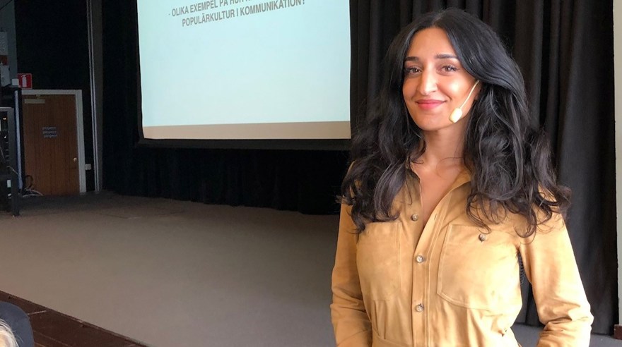 kvinna mörks långt hår. Senapsgul byxdräkt. Står framför en filmduk med en presentation på. Ser ut som en föreläsningsscen