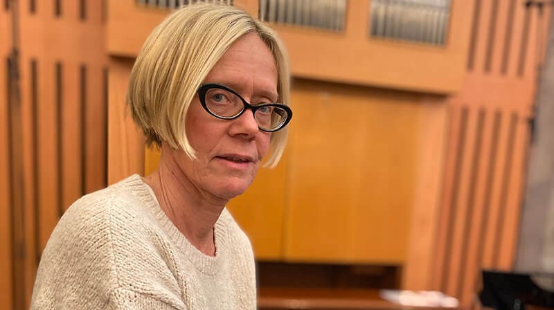 Kvinna, medelålders, kort page, blont hår, svarta glasögonbågar. Vit stickad tröja