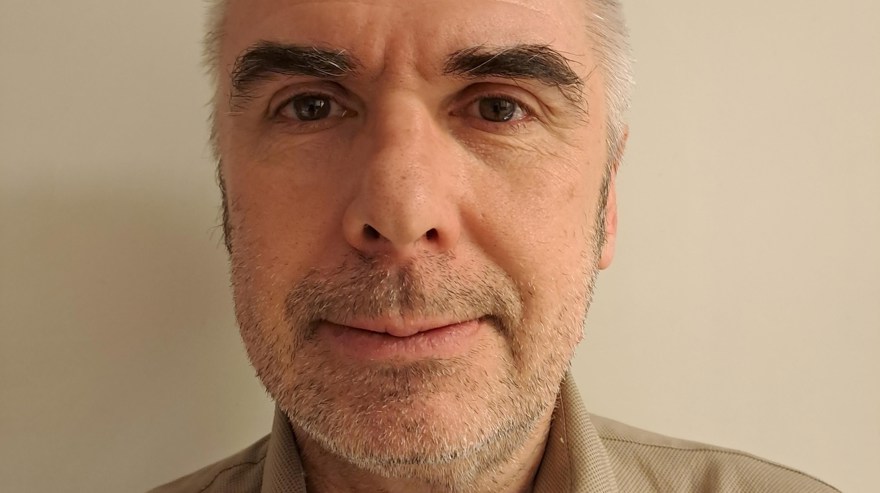 Medelålders man, mörka ögonbryn, grått skäggstubb, beige skjortkrage sticker upp i bildens nederkant