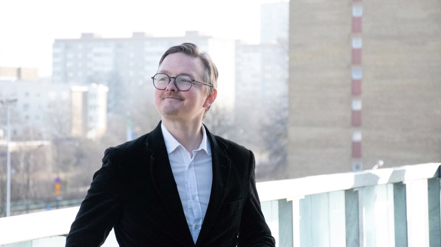 Man, runt 45 år. Kort, mellanblont hår. Lite skägg, liten mustasch. Glasögon med svarta bågar. VIt skjorta och svart kavaj. Höga hus i bakgrunden