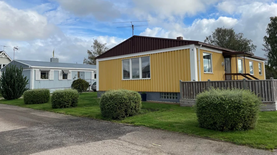 Ett par låga enplanshus, det främre gult och det bakre ljust gråblått. Rundklippta buskar framför husen. Bakom syns den blå himlen med vita molntussar.