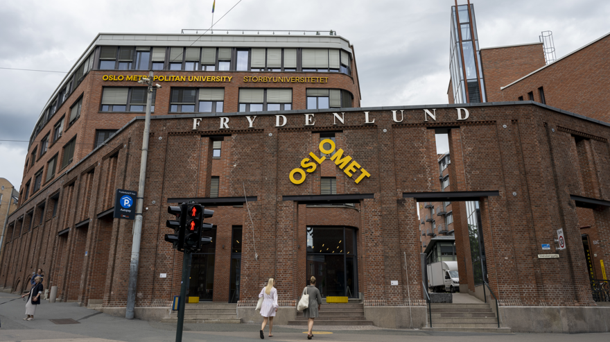 Två sommarklädda kvinnor är på väg mot en entré på en byggnad. Byggnaden är byggd i rött tegel och ovanför entrén står det med gula stora bokstäver OSLOMET. På en högre byggnad längre bort står det med gula bokstäver OSLO METROPOLITAN UNIVERSITY STORBYUNIVERSITETET.