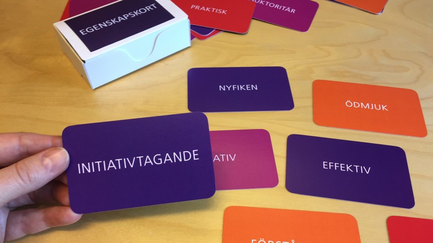 En ljus bordsskiva i trä där det ligga utspridda kort i olika röda och lila färger. På korten står olika egenskaper, som initiativtagande, nyfiken, ödmjuk och effektiv. En hand håller i det lila kortet med egenskapen initiativtagande på. Intill ligger en vit ask med fler egenskapskort.