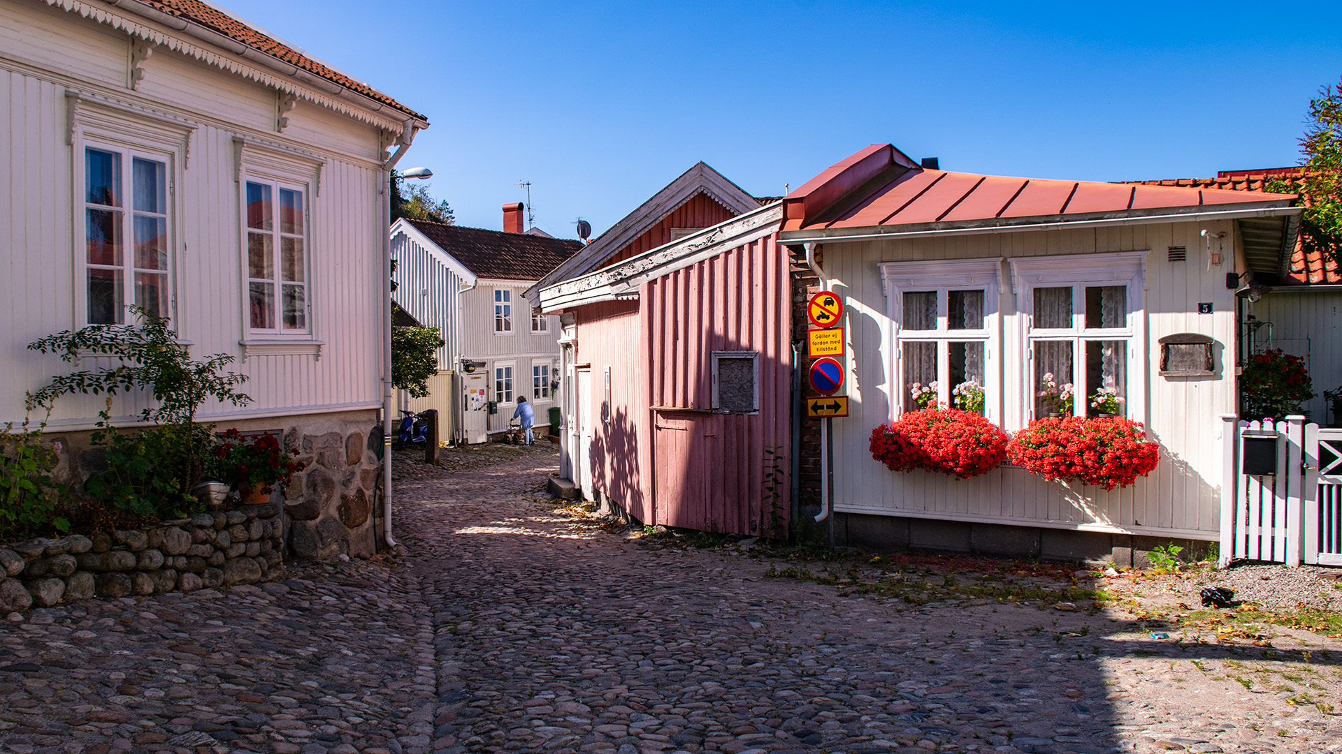 En gata med kullersten och låga äldre trähus. En kvinna med rullator står utanför ett av husen, ett annat hus har stora röda pelargoner i blomlådor vid fönstren. Solen skiner.
