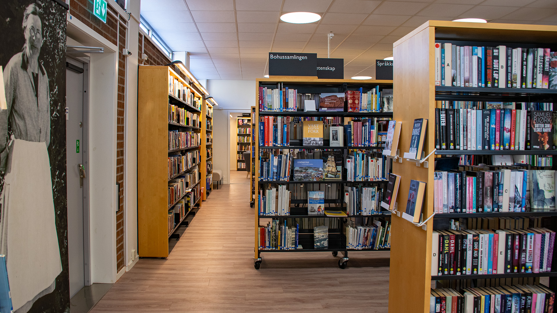 Bokhyllor i ljust trä fyllda med böcker står i biblioteksrummet. Väggen är en tegelvägg, en stor svartvit bild av en äldre kvinna i vitt förkläde och runda glasögon pryder väggen. 