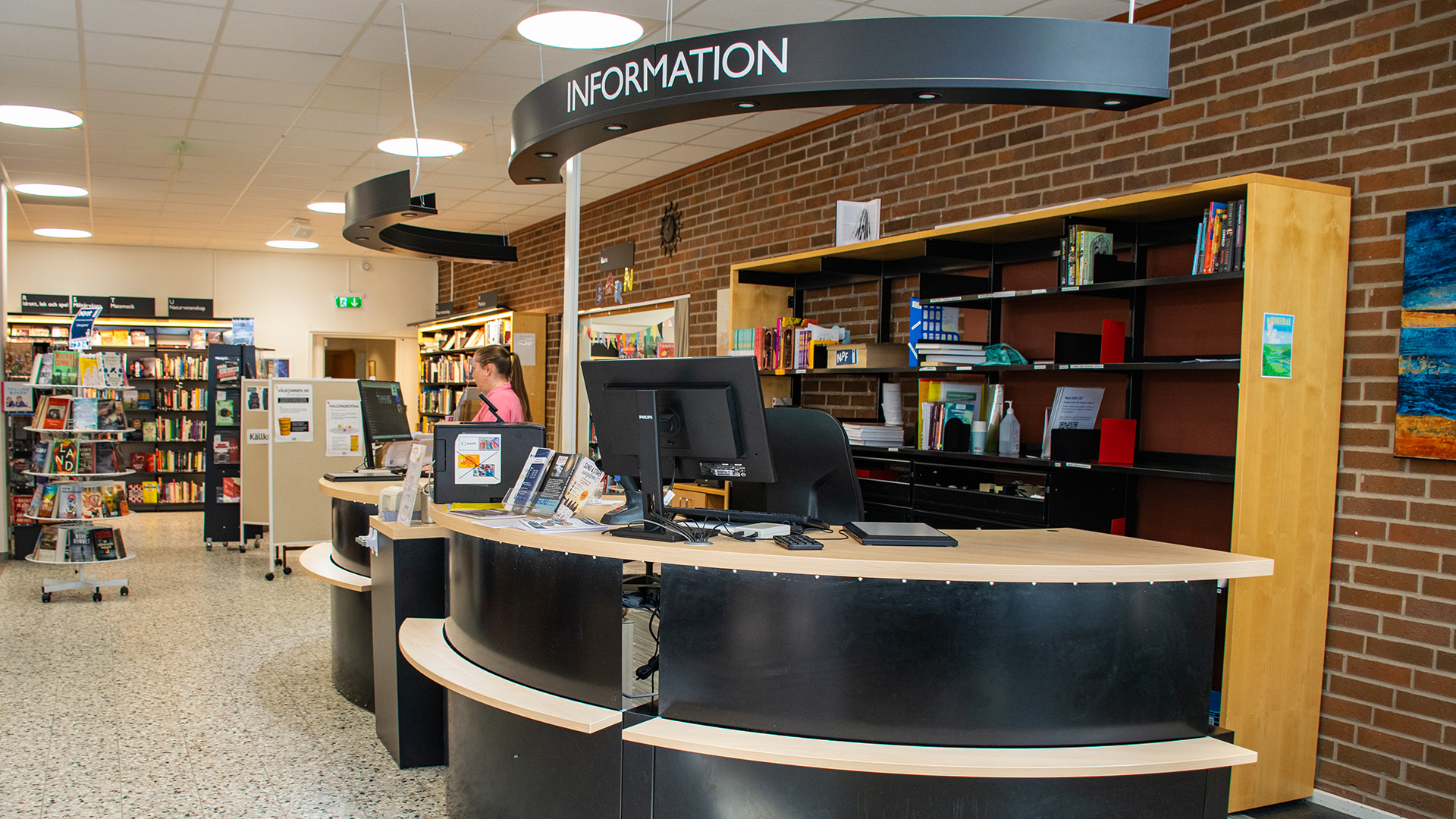 Informationsdisken på stadsbiblioteket i Strömstad är välvd och rundad i svart och ljust trä. På disken står skärmar och skyltar. Väggen bakom är i rött tegel liksom utsidan på huset. 