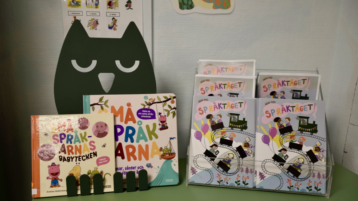 Broschyrer om Språktåget står på ett grönt bord, bakom dem tittar en uggla fram. 