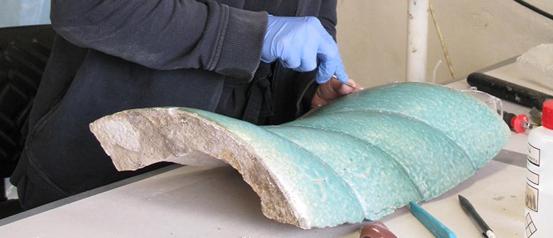 En stor bit blåturkos stengods ligger på ett bord. Med ett litet verktyg arbetar en person med något på stengodsbiten. Bara händerna syyns. Framför stengodsbiten ligger fler verktyg på bordet.