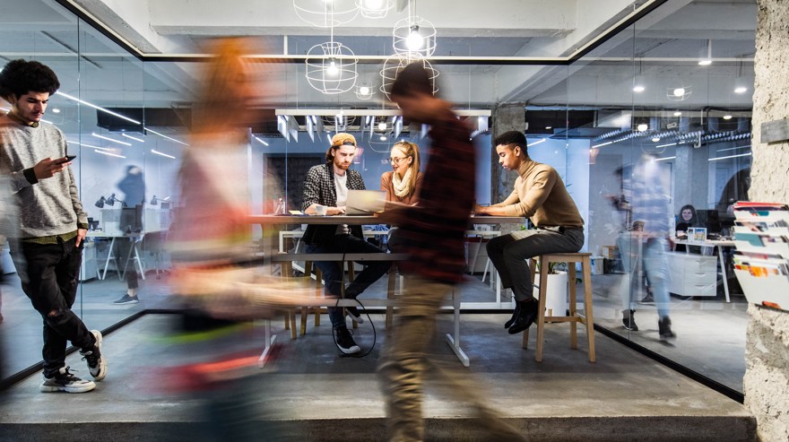 en kvinna och två män sitter vi ett högt bord och arbetar tillsammans. flera människor går förbi i förgrunden.