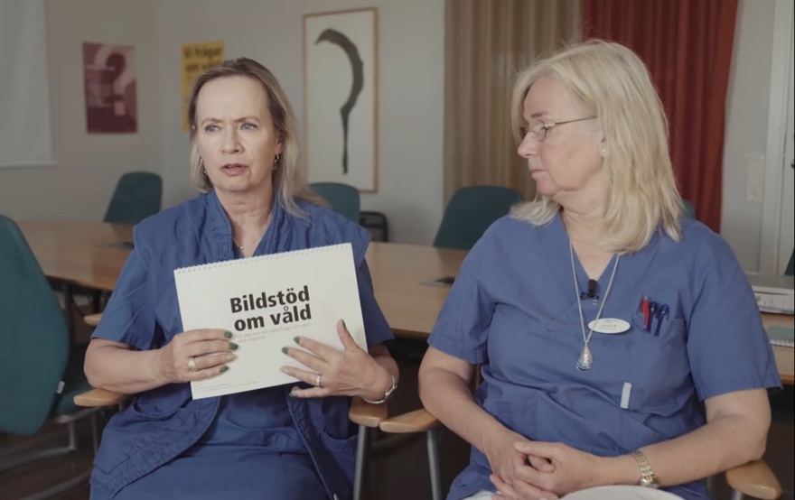 Två kvinnor iklädda blå vårdkläder. Den ena håller i ett häfte med texten "Bildstöd om våld".