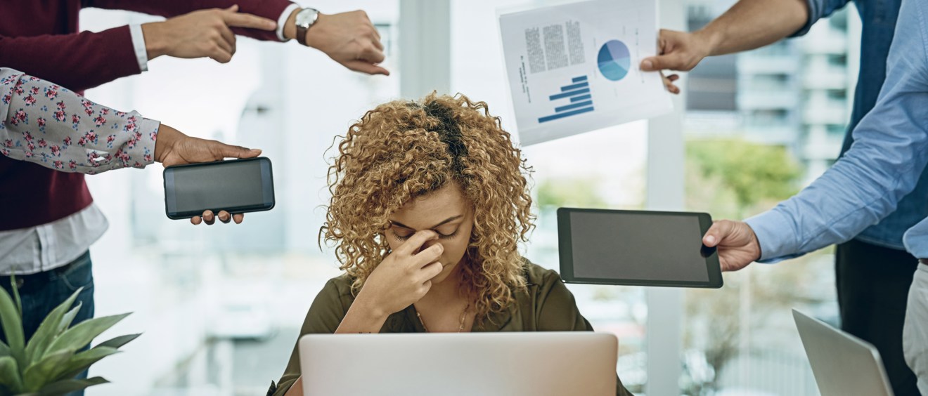 Halvbild på en person som sitter framför en dator och arbetar. Runt personen står andra personer och pekar på sitt armbandsur, håller fram en telefon, håller fram en rapport och håller fram en surfplatta. Bilden visar att personen i centrum är stressad och har hög arbetsbelastning.