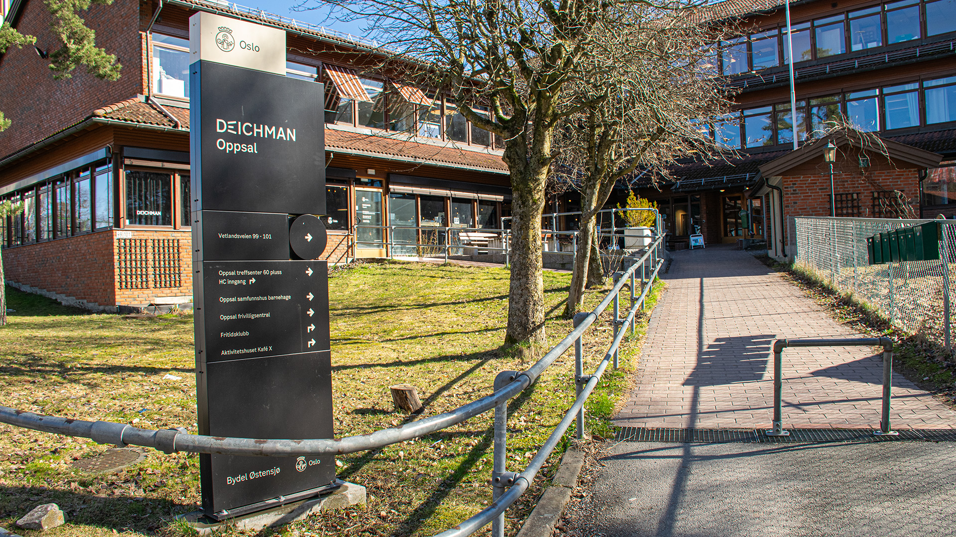 Exteriör av biblioteket och byggnaden det ligger i. En gångväg går upp till byggnaden som är i två våningar och byggd i rött tegel. Den sitter ihop med en annan trevåningsbyggnad i samma stil. Solen skiner och det är vår ute.