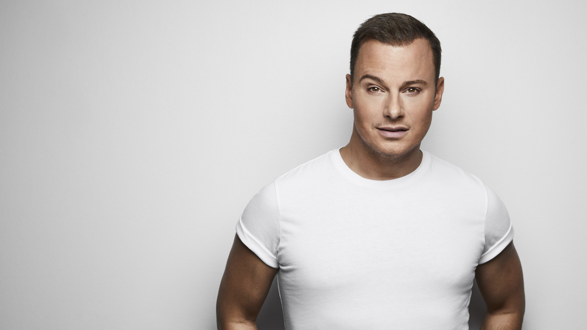  Magnus Carlsson står mot en vit vägg, han är klädd i en vit t-shirt och tittar rakt in i kameran. Magnus är i 50-årsåldern, har brunt kortklippt hår och ser solbränd ut på bilden. Han tittar med allvarlig blick mot fotografen.