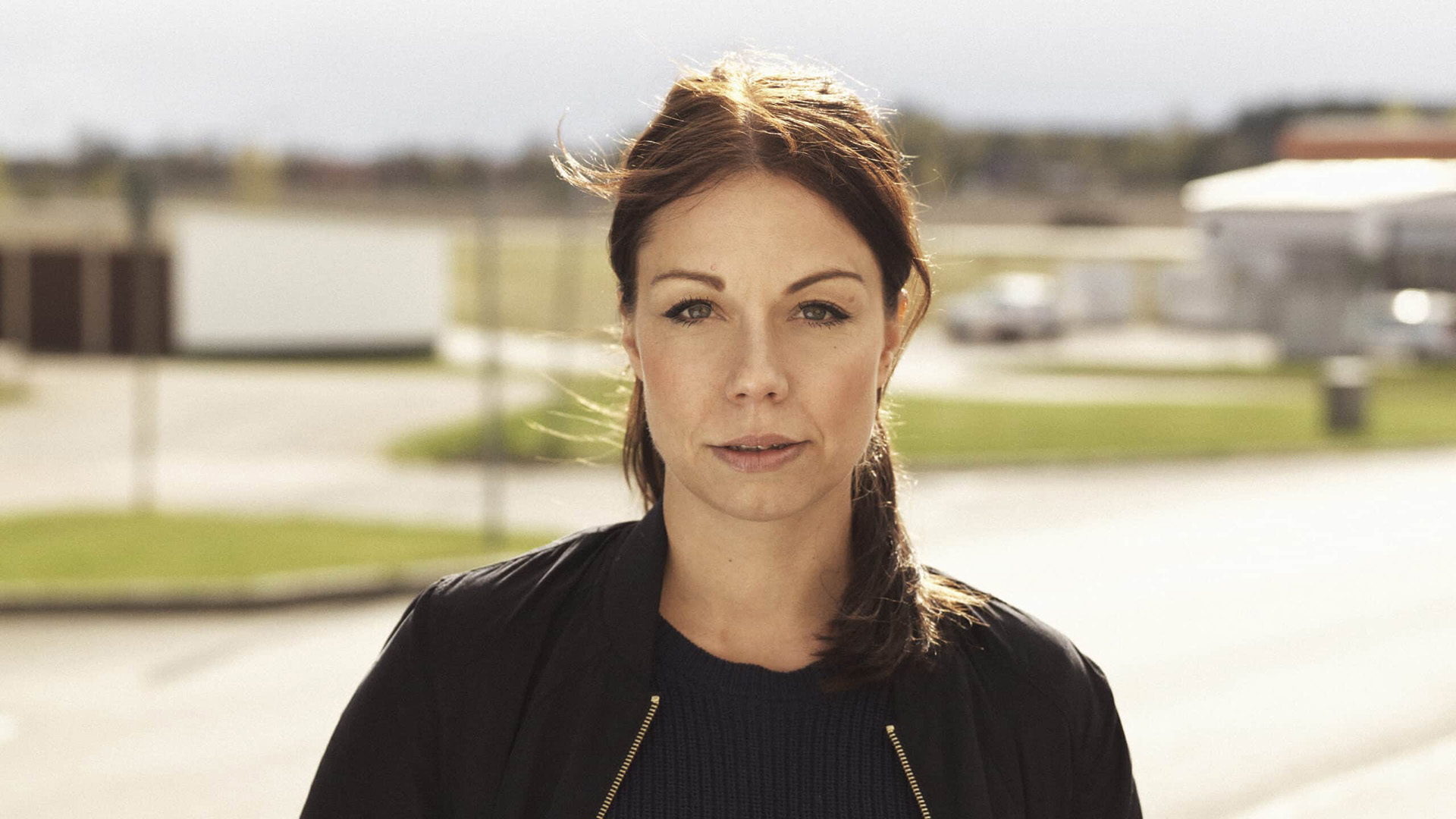 Lina Bengtsdotter är i yngre medelåldern. Hon har långt brunt hår som är samlat i en låg hästsvans. Hon står utomhus en solig dag, bakom henne finns vägar och bebyggelse. Hon är klädd i en svart jacka utan krage med en svart tröja under. Lina tittar allvarligt in i kameran. 