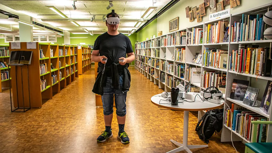 Bibliotekarie Lars Flemming testar VR inför Game On på Kinna bibliotek. Han står med VR-mask inne i biblioteksrummet, har en kontroll i varje hand. Han är klädd i jeansshorts och en svart t-shirt.