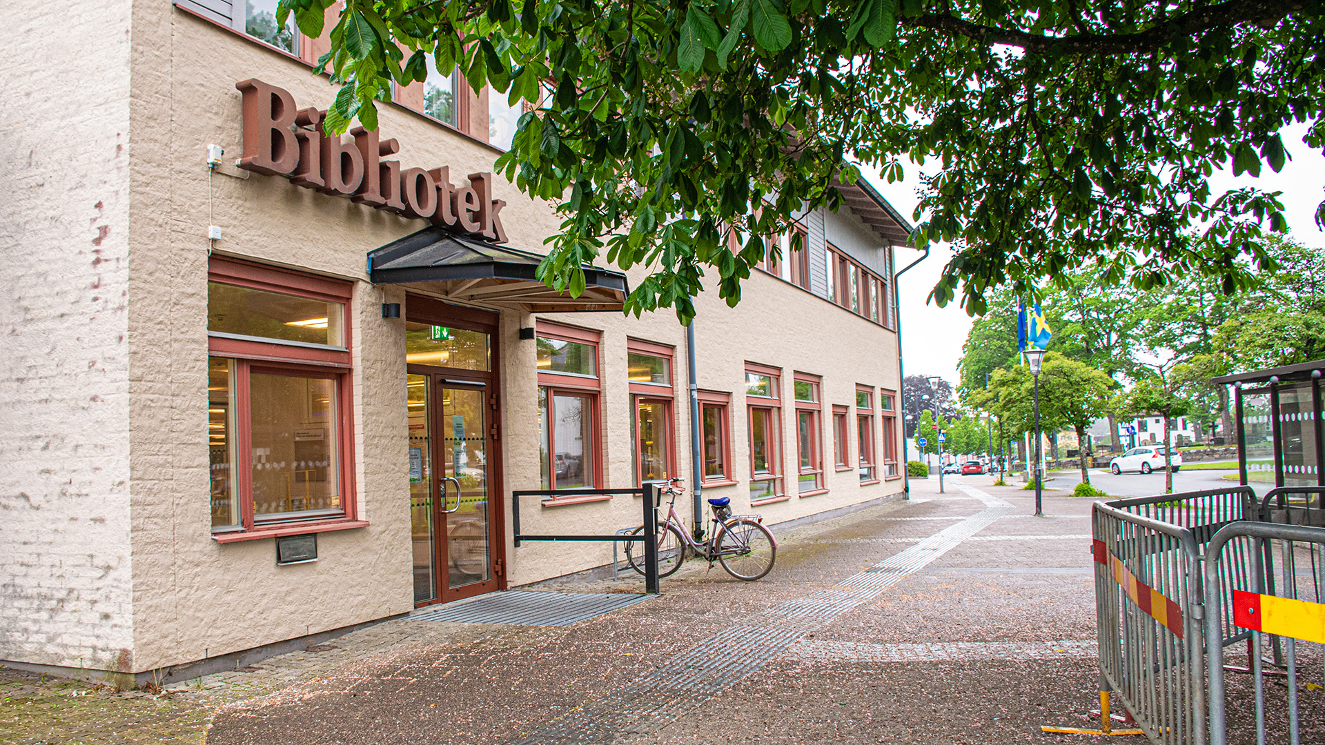 Kinna bibliotek, en byggnad i ljus tegel med texten Bibliotek ovanför glasdörren i entrén. En cykel står parkerad på gatan utanför och ett stort träd breder ut sina grenar framför huset. 