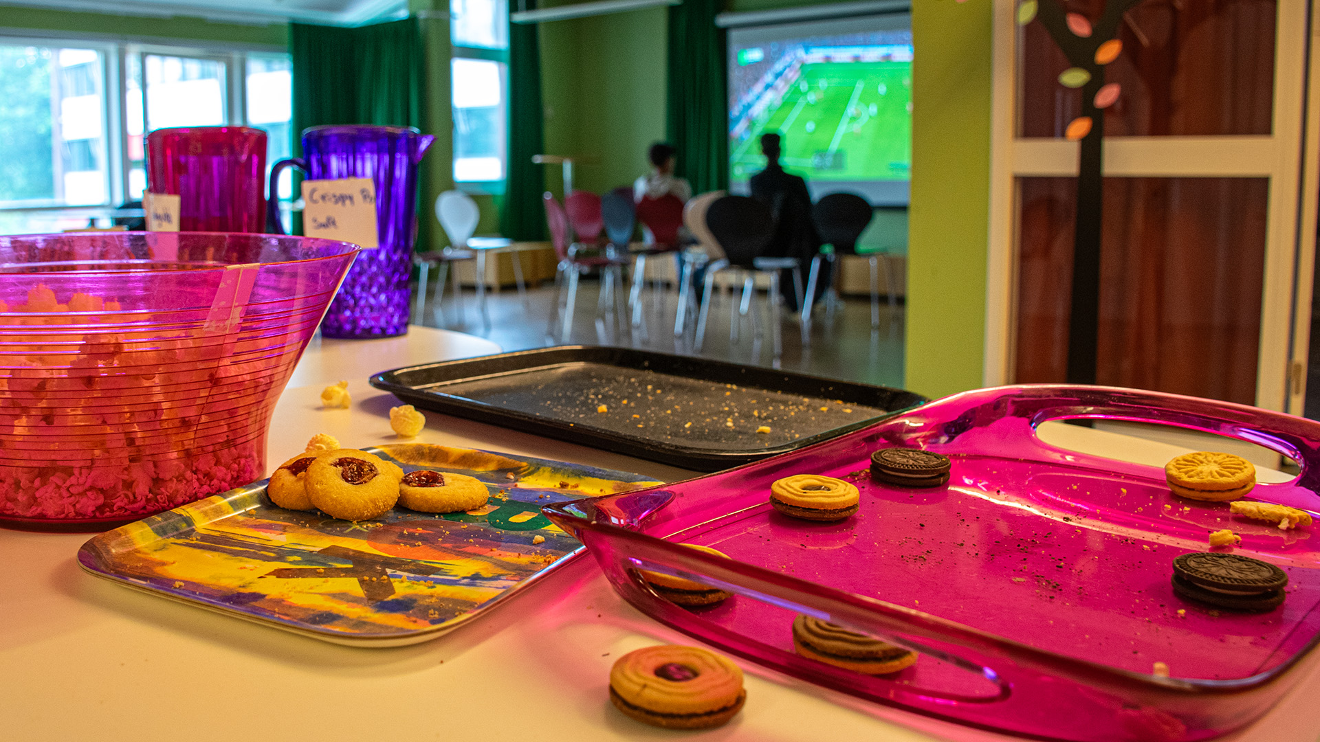Kakor och safttillbringare på ett bord. I bakgrunden spelar ett par killar FIFA med en stor filmduk framför sig. 