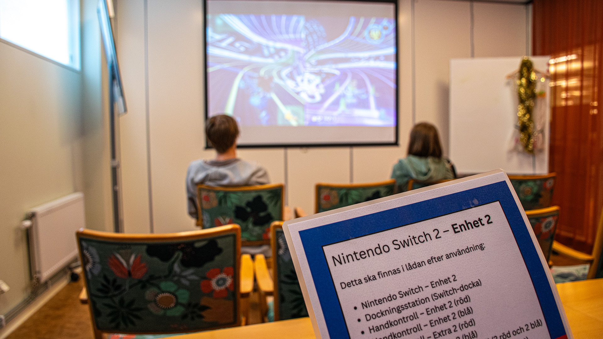 I ett mindre rum sitter en tjej och en kille i tonåren och spelar. De har blickarna på filmduken på väggen där spelet pågår. I förgrunden finns en lapp med instruktioner om Nintendo Switch. 