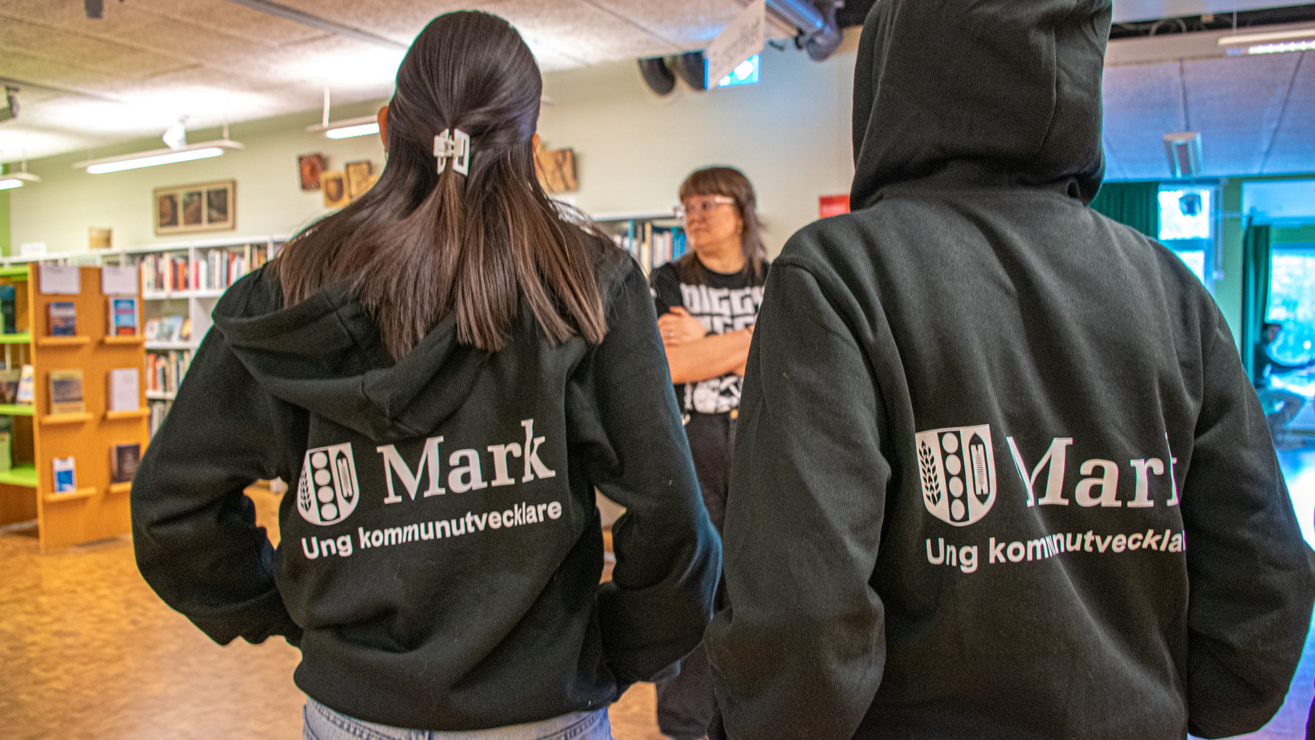 Två ungdomar klädda i svarta hoodies med texten Ung kommunutvecklare står med ryggen mot kameran. Bibliotekarie Mia-Marika Andin står framför dem och de planerar kvällen. I bakgrunden står bibliotekshyllor med böcker. 