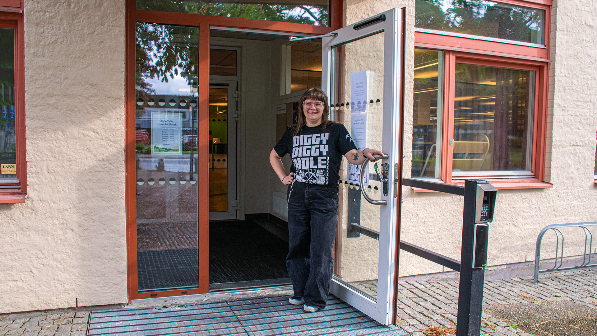 Bibliotekarie Mia-Marika Andin står i entrédörren in till biblioteket. Hon ler, hon är klädd i svarta jeans och en svart t-shirt med tryck. Hon har brunt axellångt hår och bär glasögon. 