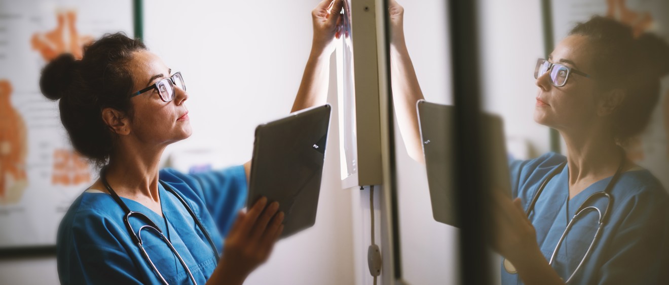 Halvbild som visar en person som har glasögon. Personen har sjukhuskläder och ett stetoskop runt halsen och tittar på röntgenbilder.