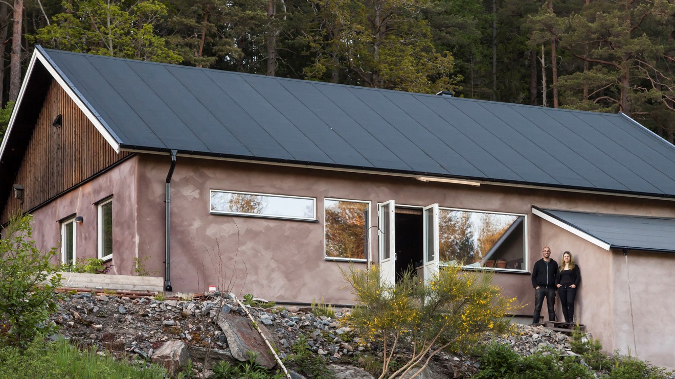 Två personer står framför ett putsat hus med papptak.